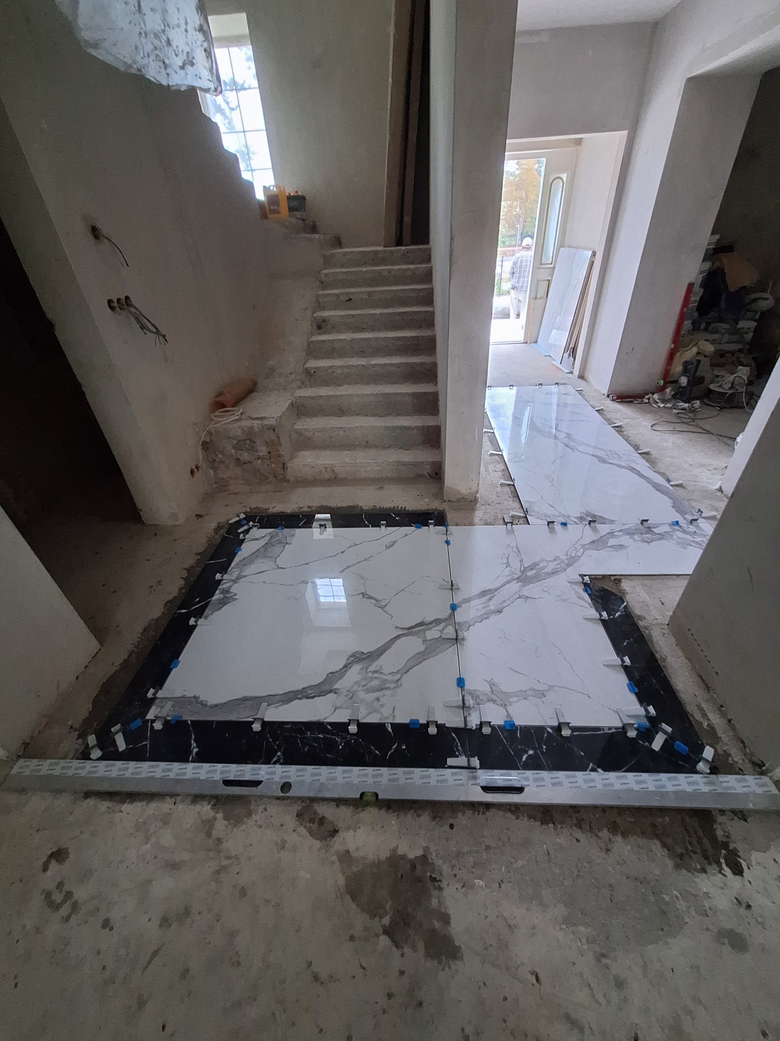 Montaż płytek imitujących marmur na podłodze w holu, widoczne krzyżyki dystansowe i poziomica, surowy stan ścian i schodów.