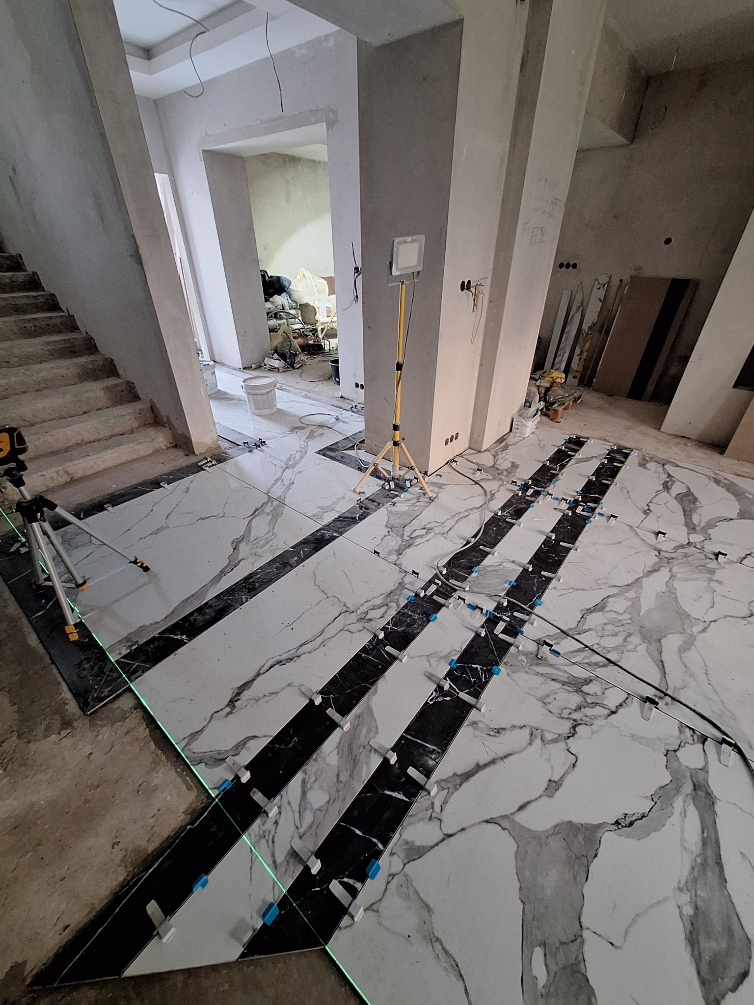 Układanie podłogi z płytek imitujących marmur w pomieszczeniu z surowymi ścianami, widoczny laser krzyżowy i system poziomowania płytek.