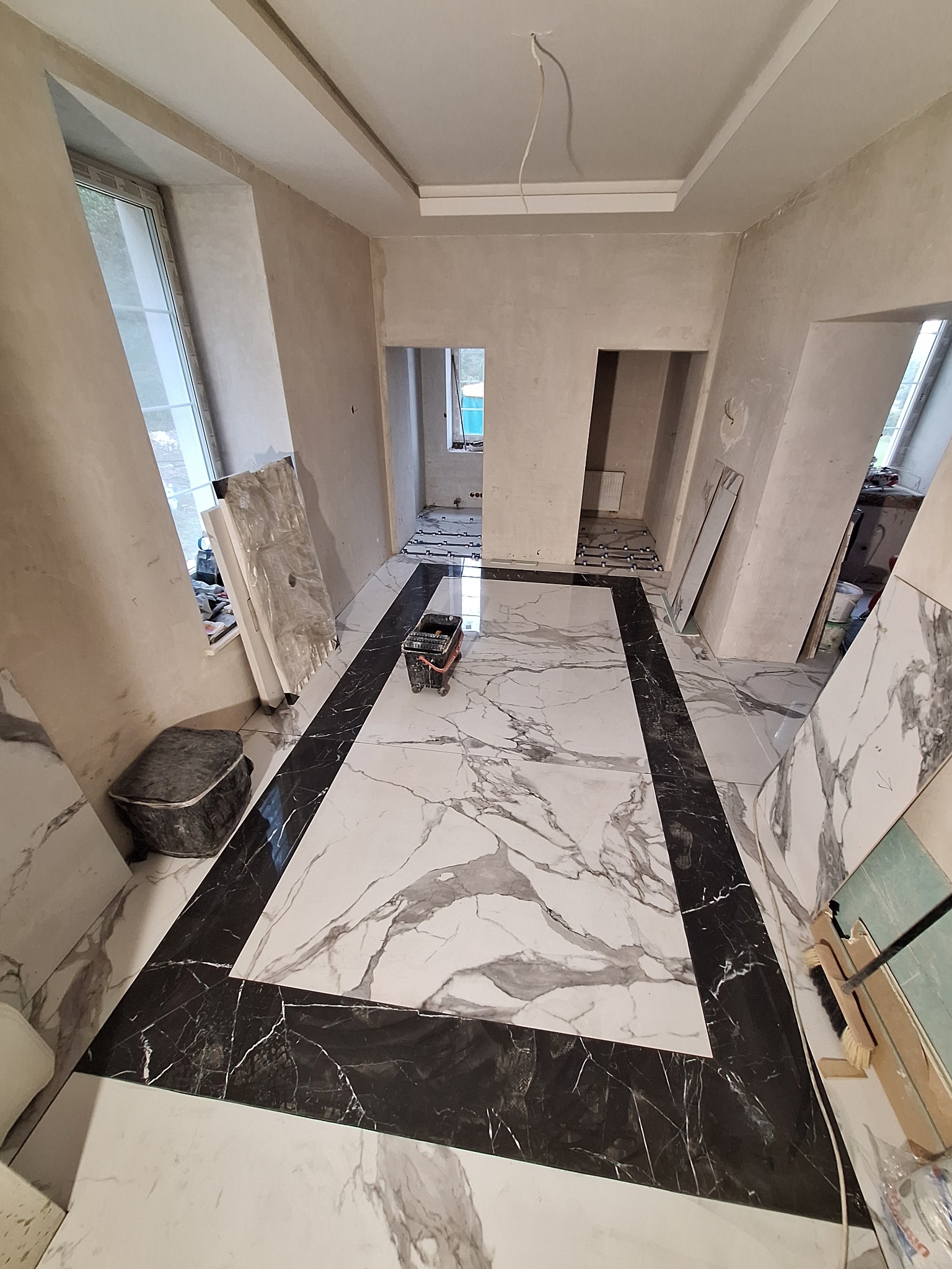 Układanie płytek podłogowych imitujących marmur w trakcie remontu; widoczny wózek do fugowania, płyty do docinania i surowe ściany.