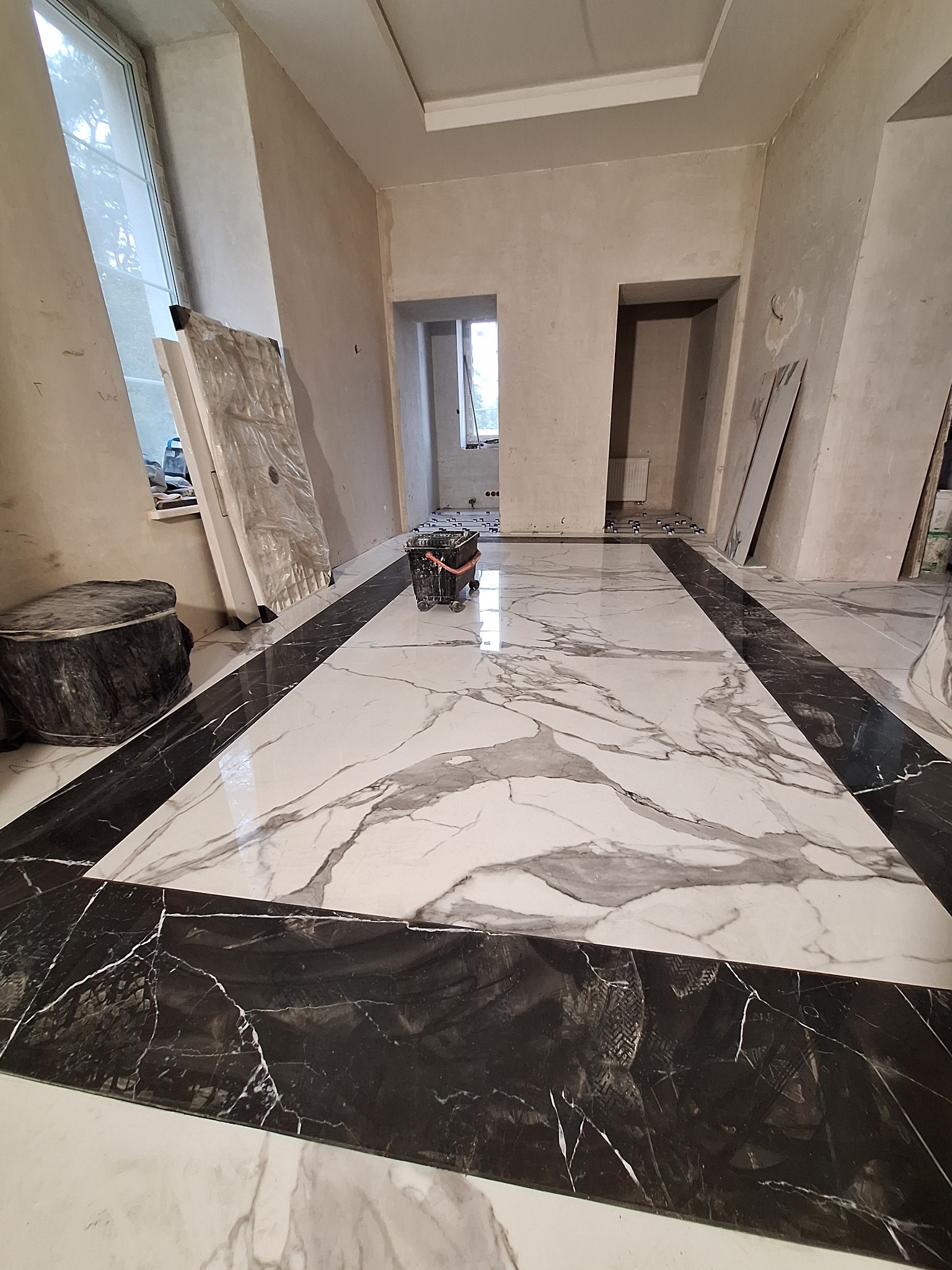 Ułożona podłoga z płytek imitujących marmur z czarną obwódką w surowym wnętrzu, widoczne ślady butów na obwódce i wiadro na kółkach.