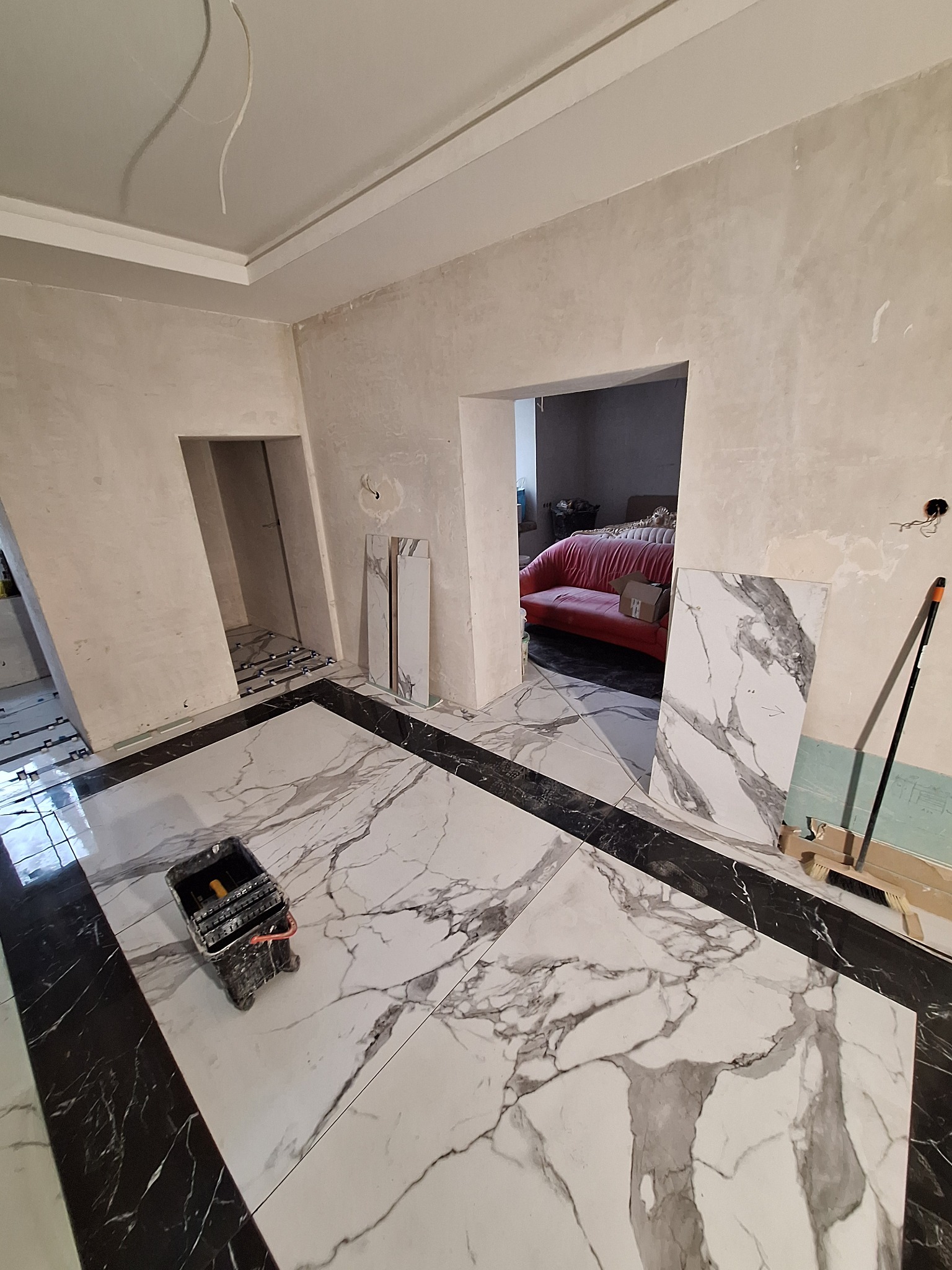 Wnętrze w trakcie prac, widoczne płytki imitujące marmur na podłodze z czarnymi obramowaniami, narzędzia glazurnicze, oraz przygotowane do montażu płytki przy ścianie.