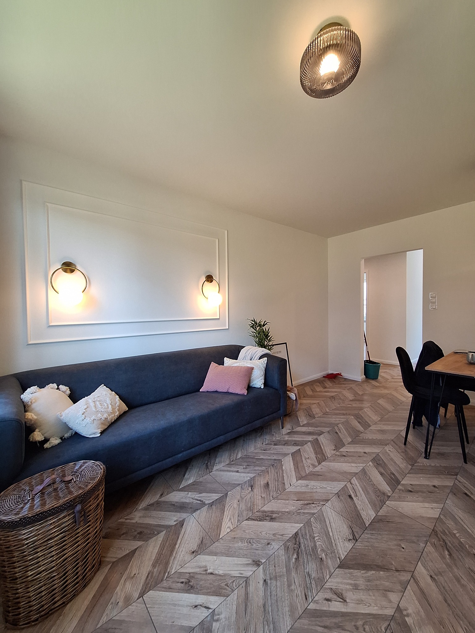 Salon z ciemnoszarą sofą, drewnianą podłogą ułożoną w jodełkę, dekoracyjną listwą na ścianie z dwoma okrągłymi lampami, wiklinowym koszem i widokiem na jadalnię.