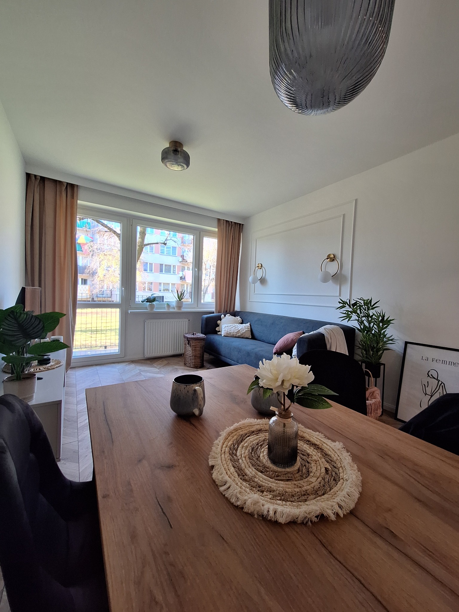 Jasne wnętrze salonu z drewnianym stołem, szarą sofą i dekoracyjną sztukaterią na ścianie. Na stole wazon z kwiatami i podkładka z frędzlami. Widok na budynek mieszkalny za oknem.