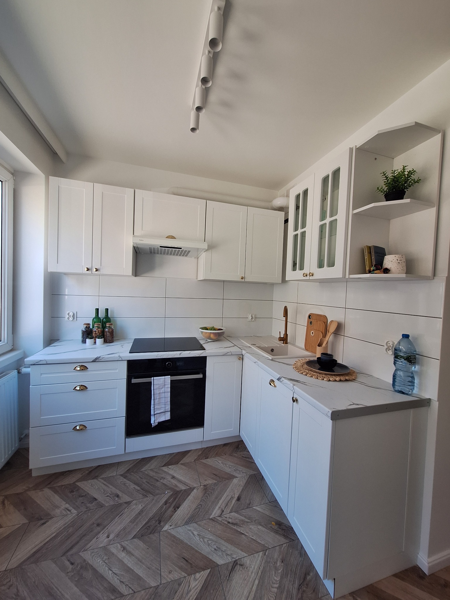 Jasna kuchnia z białymi szafkami i blatem imitującym marmur, złota armatura, parkiet w jodełkę, nowoczesne oświetlenie sufitowe.