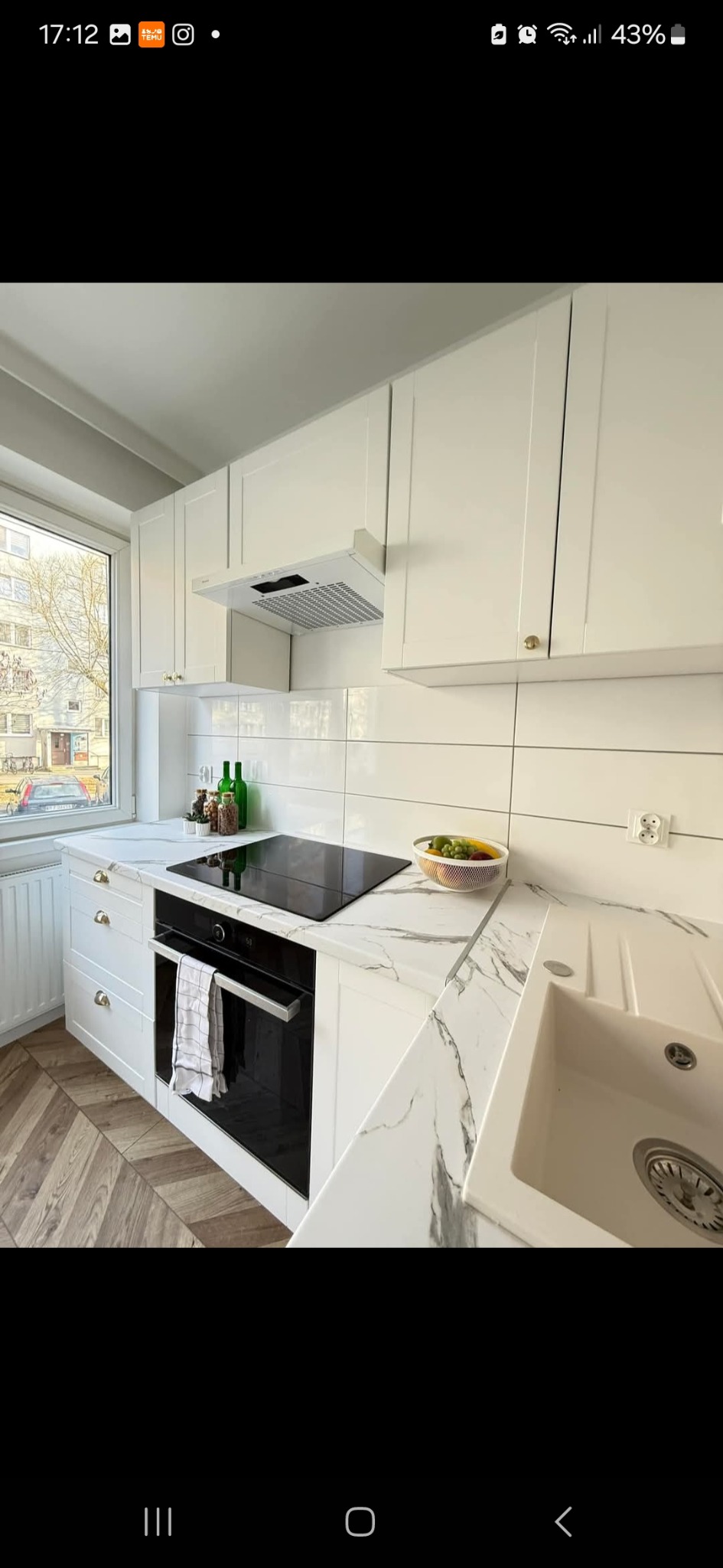 Jasna kuchnia z białymi szafkami, blatem imitującym marmur i drewnianą podłogą w jodełkę; widok na okno z zabudową blokową na zewnątrz.