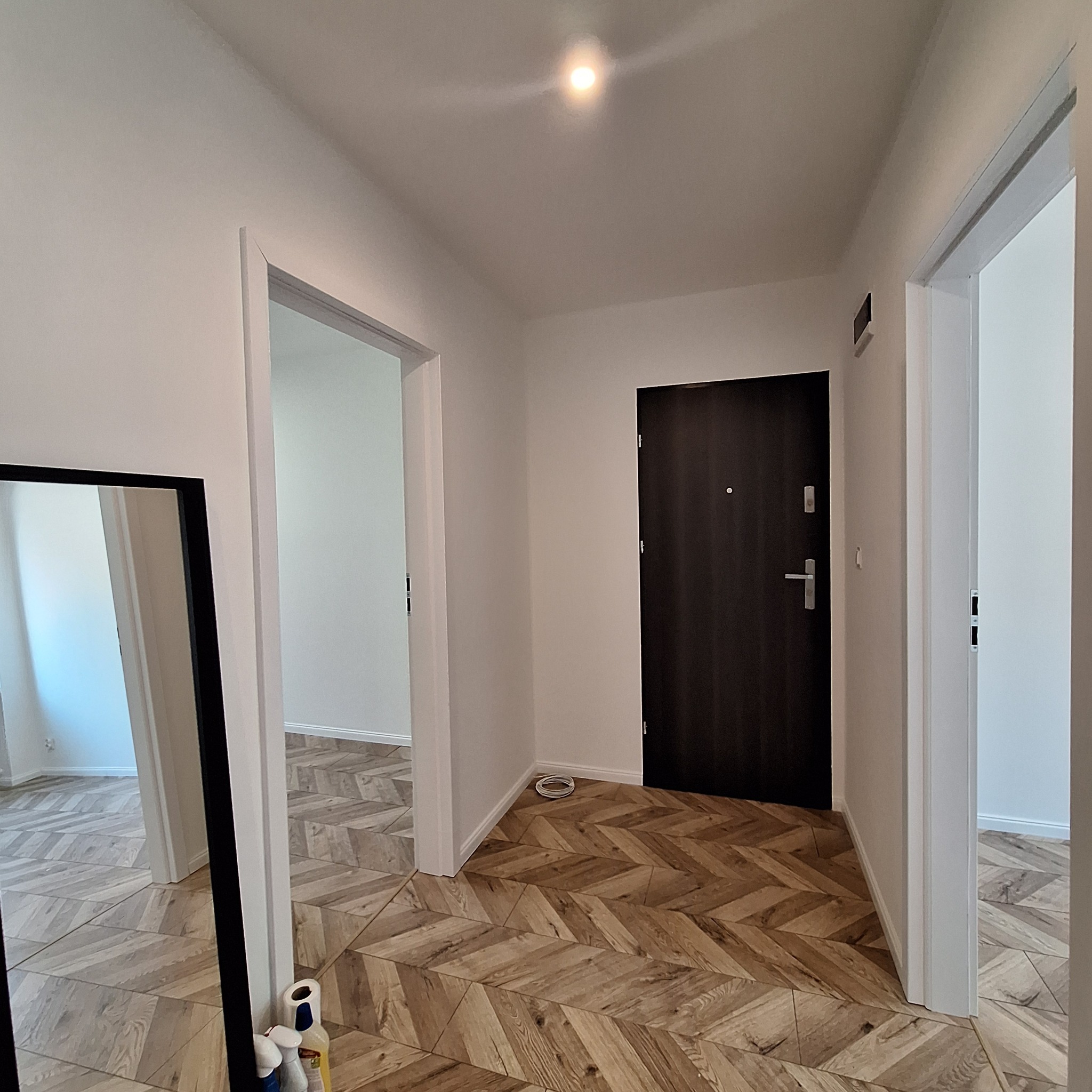 Korytarz po remoncie z podłogą ułożoną w jodełkę z drewnopodobnych paneli, białe ściany i drzwi, widoczne przejścia do innych pomieszczeń, w rogu przy lustrze leżą środki czystości.