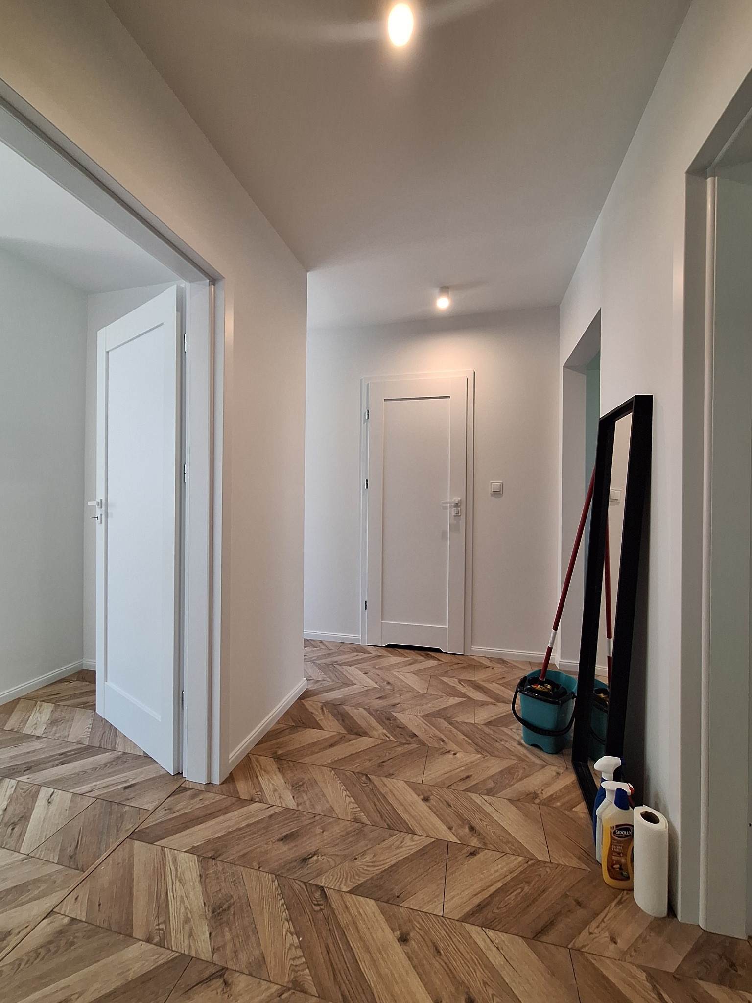Jasne wnętrze po remoncie: parkiet ułożony w jodełkę, białe ściany i drzwi, widoczne akcesoria do sprzątania.