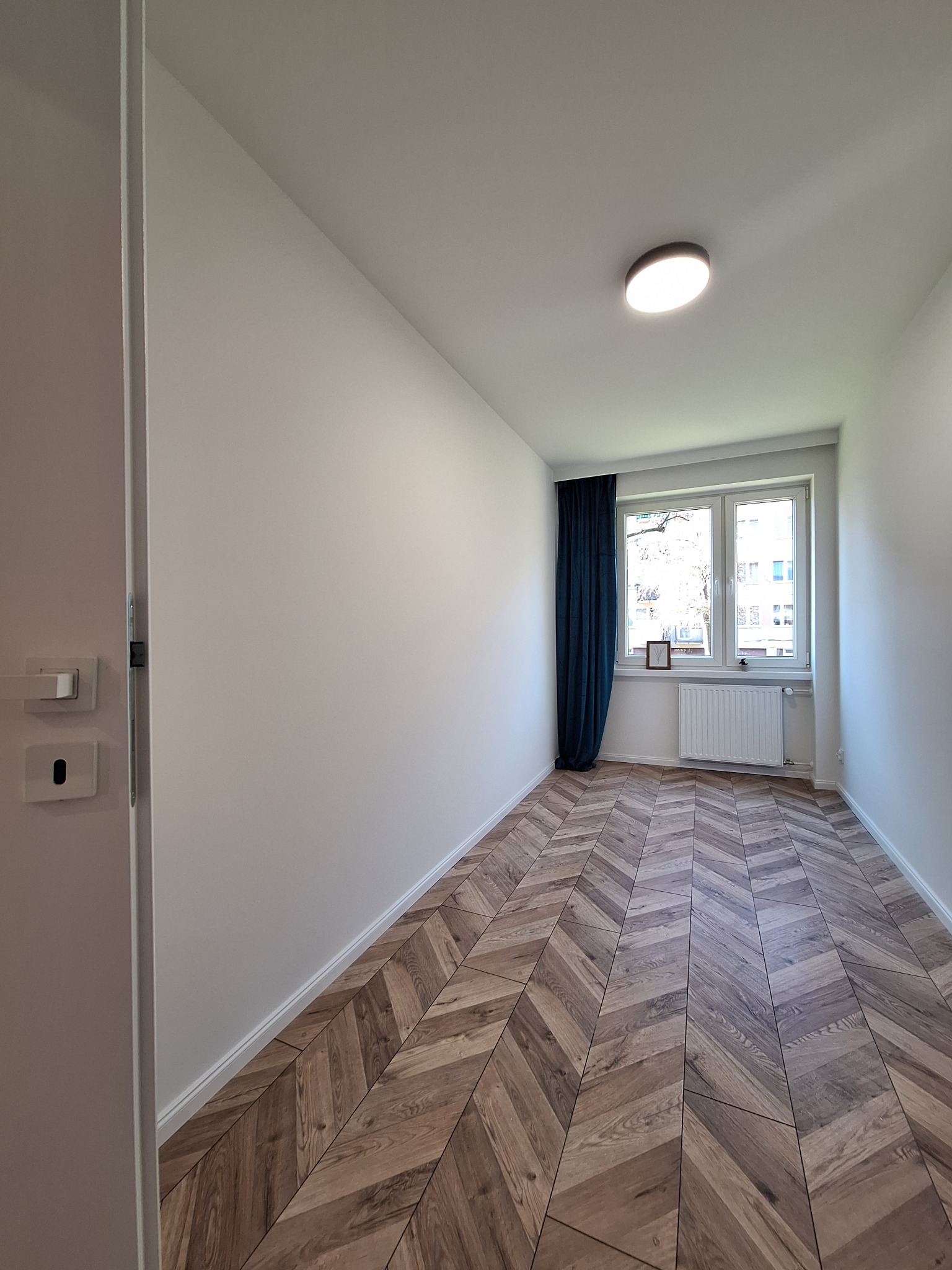 Pusty pokój po remoncie z parkietem ułożonym w jodełkę, białe ściany, okno z niebieskimi zasłonami i grzejnikiem.