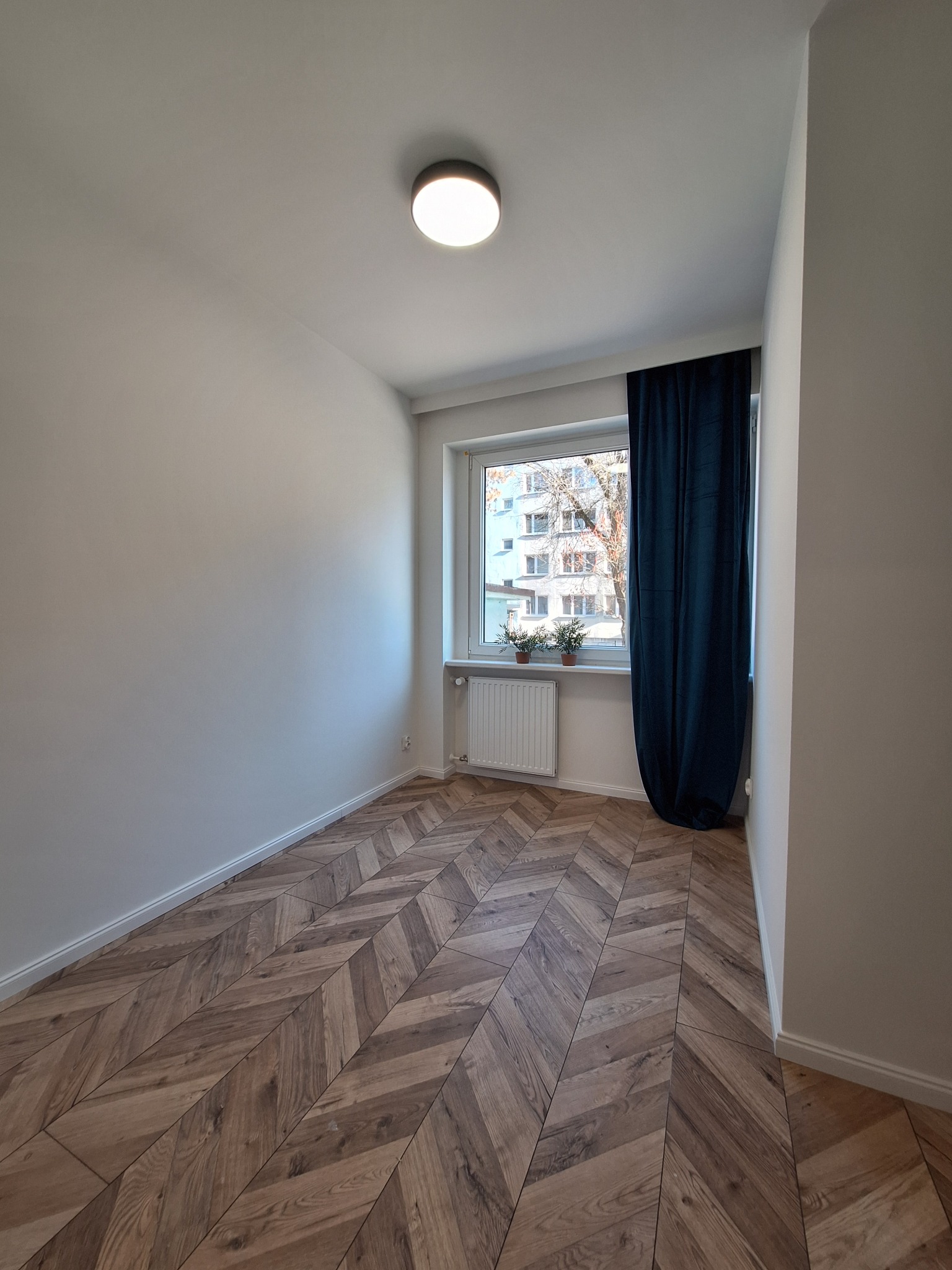 Pusty pokój po remoncie z ułożoną parkietową podłogą w jodełkę, pomalowanymi na biało ścianami i oknem z ciemnozieloną zasłoną.