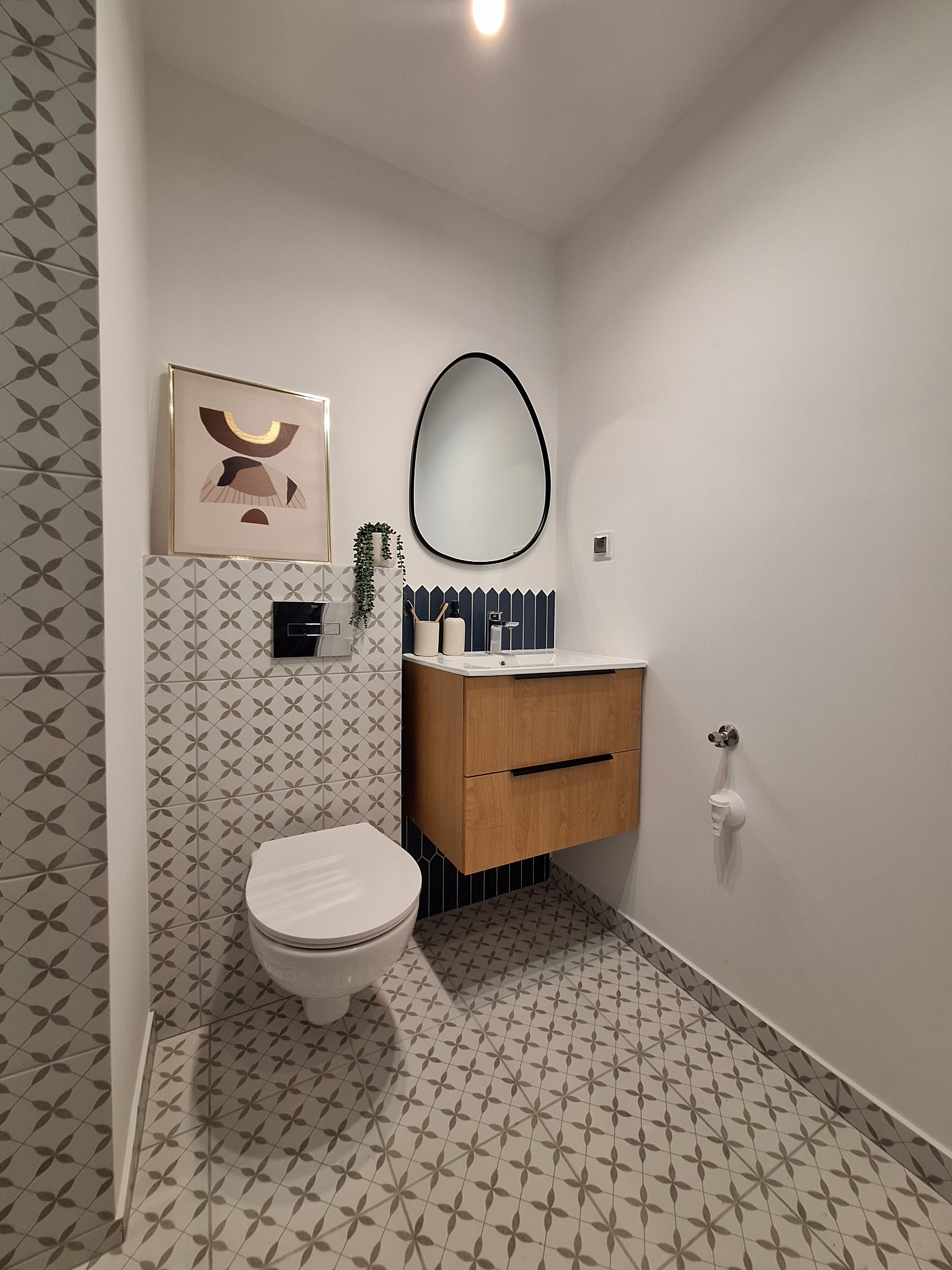 Nowoczesna łazienka z wiszącą szafką z drewnianymi frontami, owalnym lustrem w czarnej ramie, toaletą i płytkami w geometryczny wzór na podłodze i ścianie za toaletą.