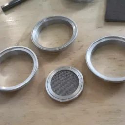 Metalowe pierścienie i okrągły element z siatką, wykonane na obrabiarce CNC, leżące na drewnianym blacie. Widoczne ślady obróbki.