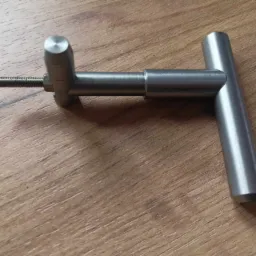 Metalowy uchwyt w kształcie litery T z wystającym gwintem, leżący na drewnianej powierzchni.