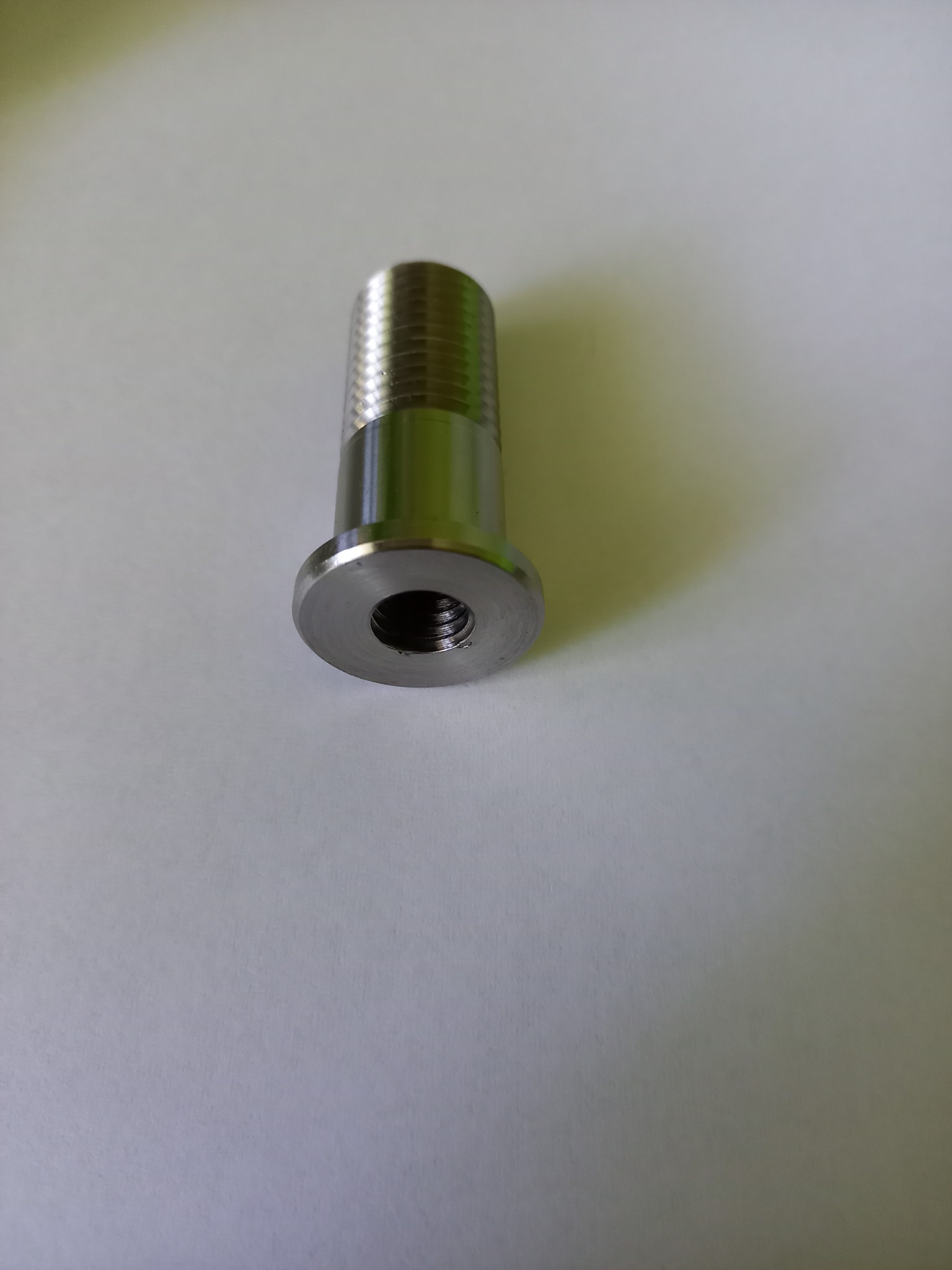 Metalowy element z gwintem zewnętrznym i wewnętrznym, widoczne ślady obróbki skrawaniem, leżący na gładkiej powierzchni.