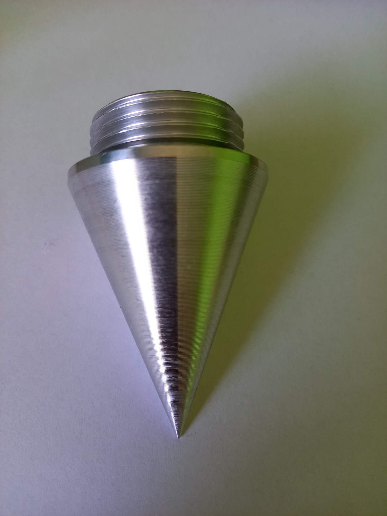 Stożkowy, metalowy element z gwintem na górze, leżący na jasnym tle. Widoczne ślady obróbki na powierzchni.