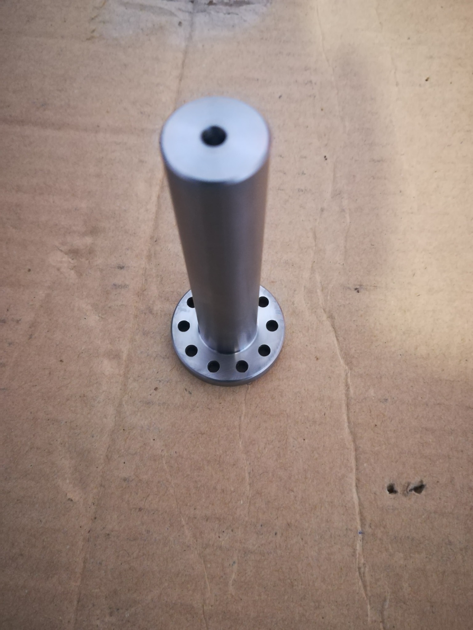 Metalowy element z kołnierzem i centralnym otworem, widoczne ślady obróbki, leżący na kartonowym tle.