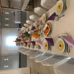 Uroczysty stół w Pacanowie, nakryty białym obrusem, z białymi krzesłami, talerzami z spaghetti, fioletowymi serwetkami, szklankami i dzbankami z sokiem, przygotowany na przyjęcie.