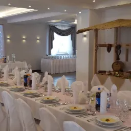 Uroczysta sala bankietowa z długim stołem nakrytym białymi obrusami i krzesłami w pokrowcach, dekoracje z serduszkami i światełkami w tle, oraz stoisko z wędlinami i beczką po prawej stronie.