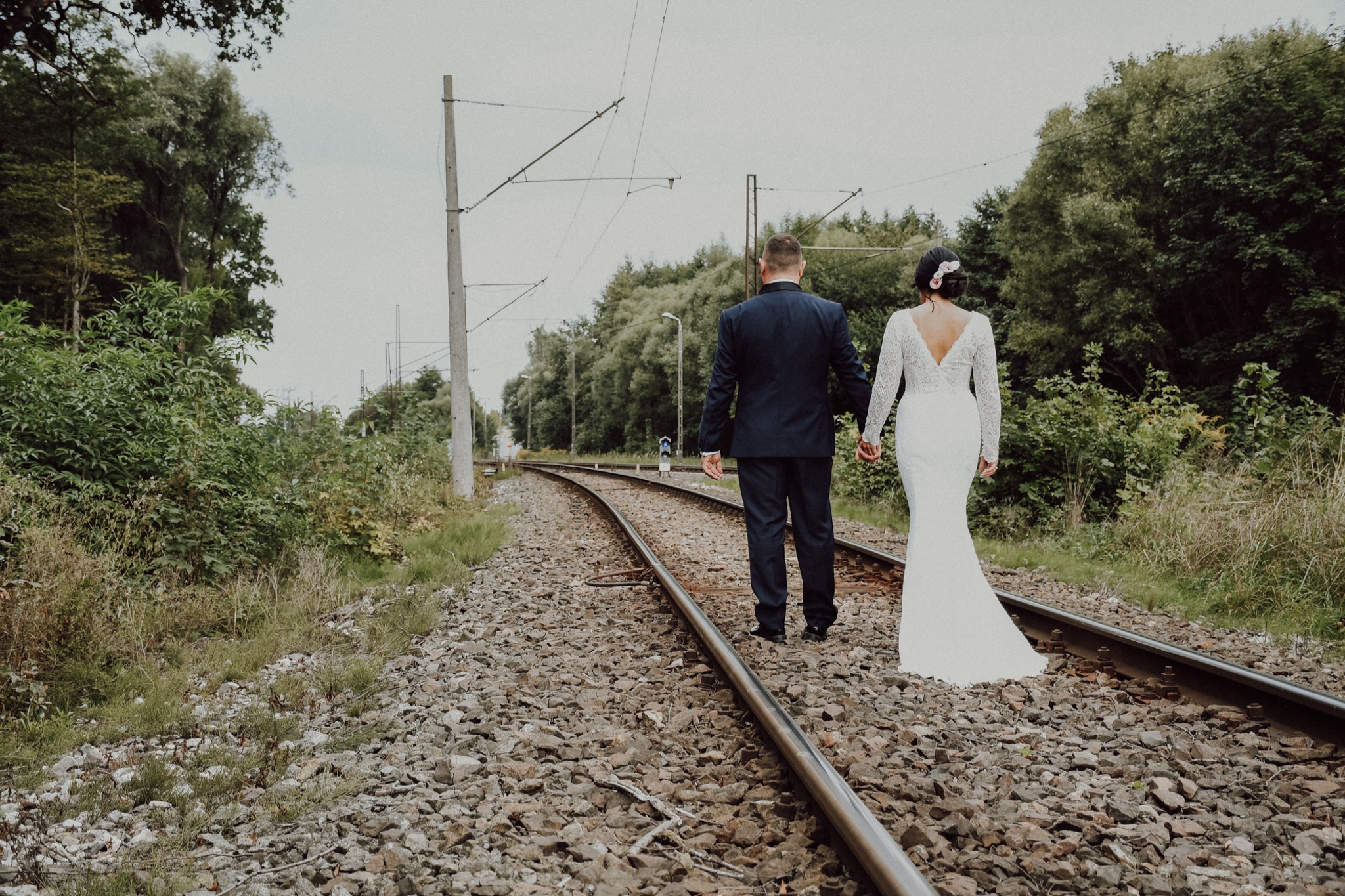 Para młoda, on w granatowym garniturze, ona w koronkowej sukni z długim rękawem, idą trzymając się za ręce po torach kolejowych, z tyłu widok na drzewa i słupy trakcji elektrycznej.