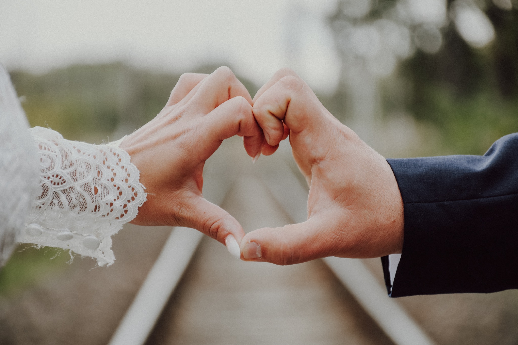 Dłonie nowożeńców formują serce na tle torów kolejowych, ona w koronkowej sukni, on w ciemnym garniturze.