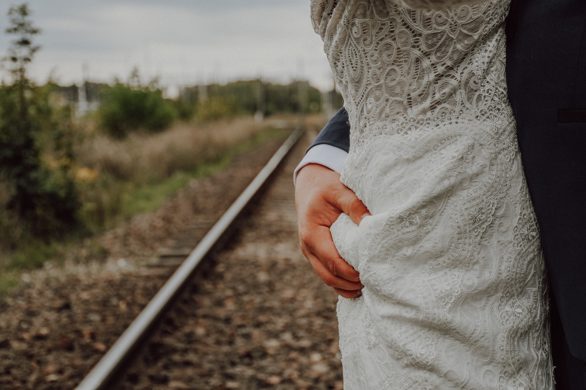 Mężczyzna w granatowym garniturze obejmuje kobietę w koronkowej sukni ślubnej na tle torów kolejowych i zieleni.