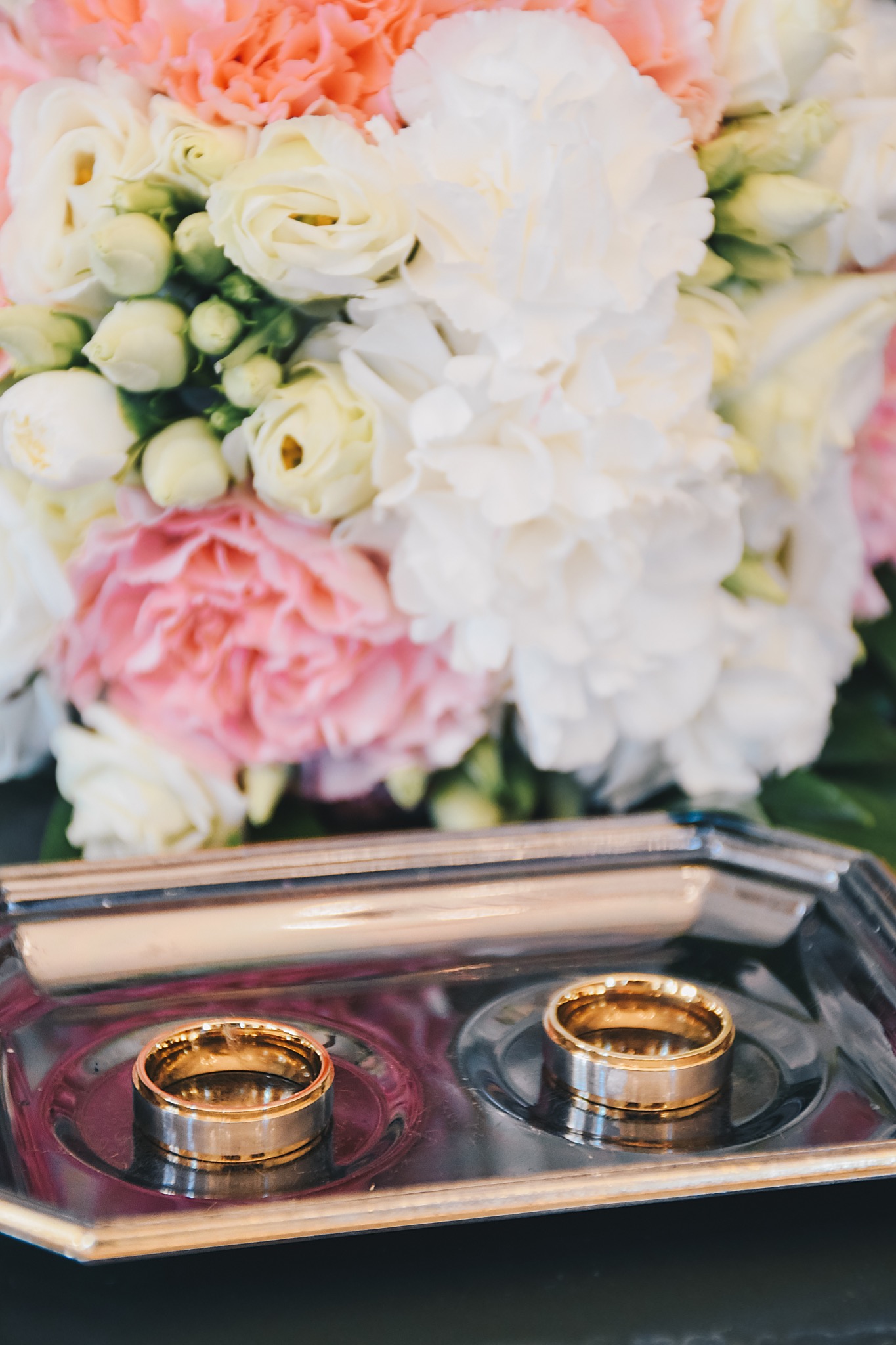 Dwa obrączki ślubne dwukolorowe (złoto i srebro) leżące na srebrnej tacy, w tle bukiet kwiatów w odcieniach bieli, różu i zieleni.