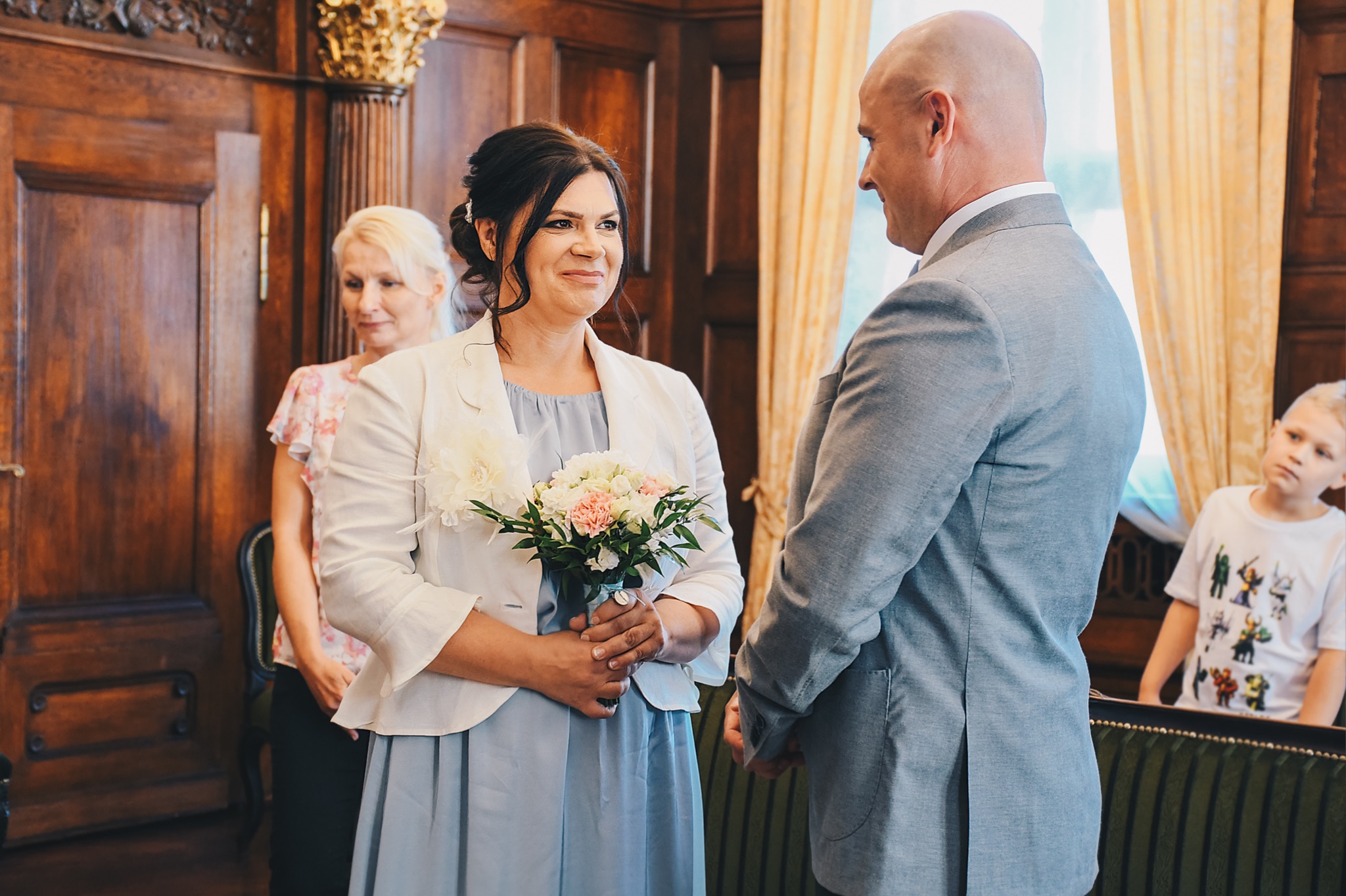 Panna młoda w jasnoniebieskiej sukience i białym żakiecie, trzyma bukiet ślubny z różowych i białych kwiatów, uśmiecha się do pana młodego w szarym garniturze, w tle drewniane drzwi i obserwujący...