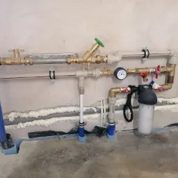 Nowa instalacja wodna z licznikiem, filtrem i zaworami, zamontowana przy ścianie z widocznymi śladami pianki montażowej i izolacji podłogowej.