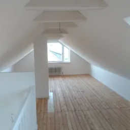 Poddasze z drewnianą podłogą i białymi ścianami, widoczne trapezowe panele sufitowe, okno z grzejnikiem oraz fragment białej balustrady.