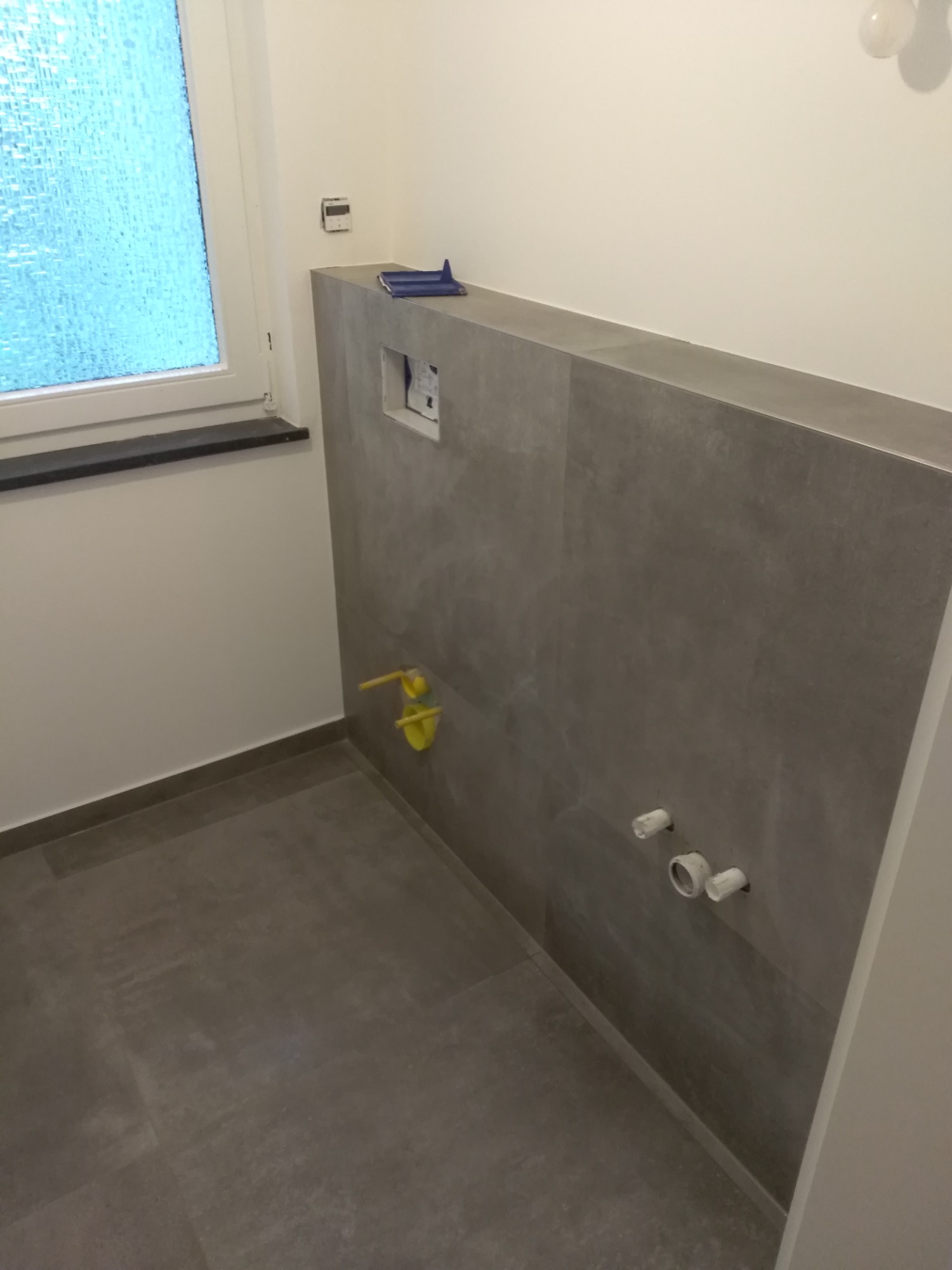 Szara łazienka w trakcie remontu: położona glazura na ścianie, widoczne rury i instalacja podtynkowa, okno z ozdobną szybą.