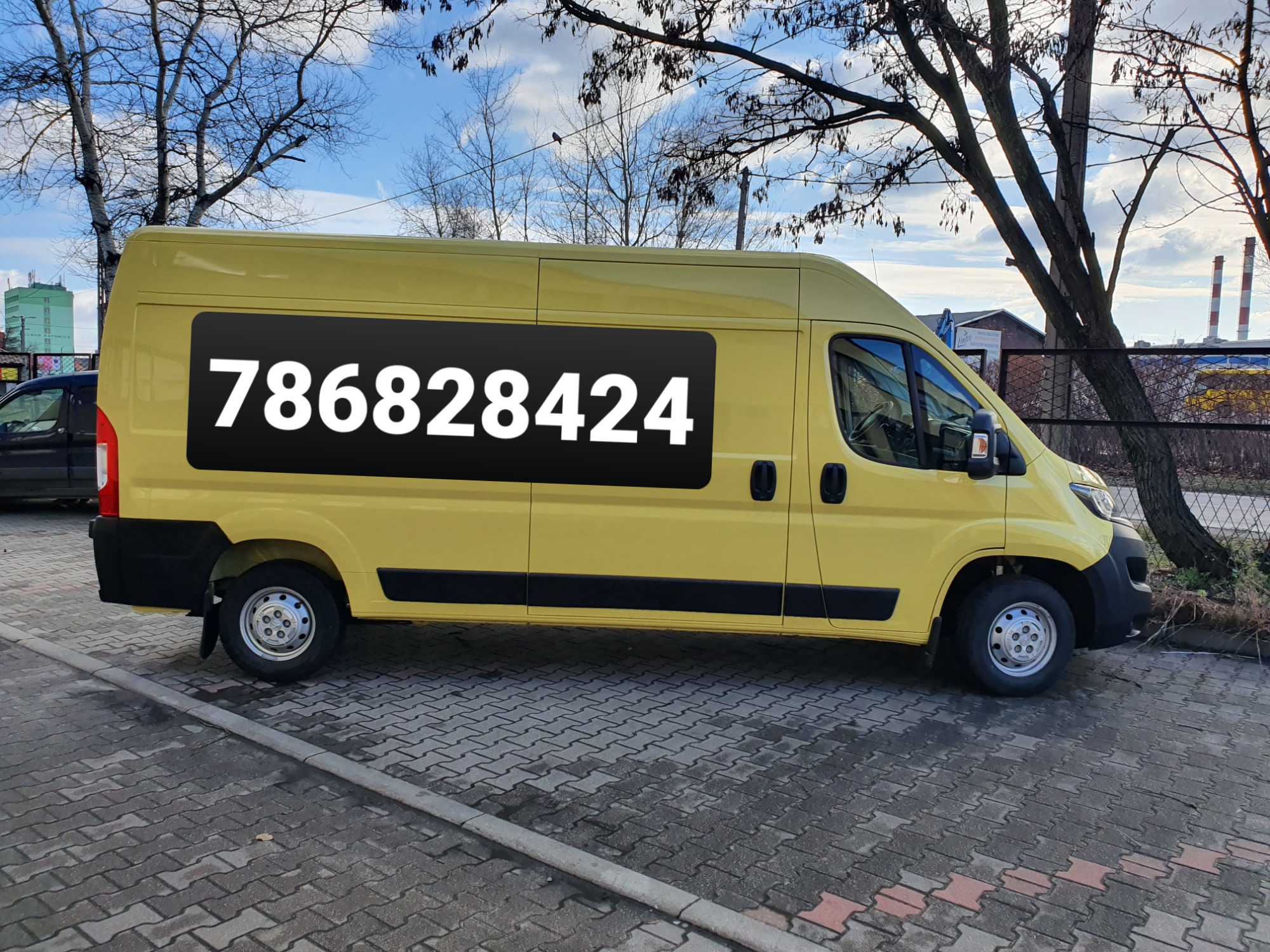 Żółty bus dostawczy z dużym numerem telefonu na boku, zaparkowany na chodniku z kostki brukowej, w tle drzewa bez liści i budynki przemysłowe.