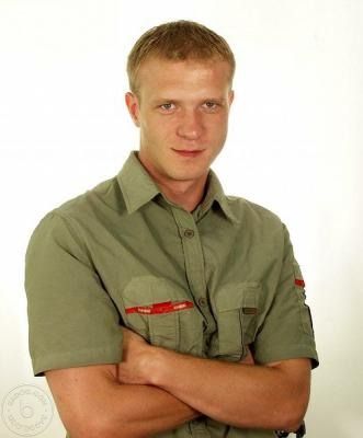 Mężczyzna o jasnych włosach w zielonej koszuli z krótkim rękawem, skrzyżowanymi rękami na białym tle, spogląda w obiektyw z lekkim uśmiechem.