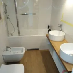 Nowoczesna łazienka we Wrocławiu z dwiema okrągłymi umywalkami na drewnianym blacie, wiszącym bidetem i toaletą oraz kabiną prysznicową z wzorzystą ścianą.