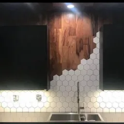 Fragment ściany w kuchni z dekoracyjnym panelem z drewna i białych, heksagonalnych płytek, częściowo ułożonych, oświetlonych od dołu.