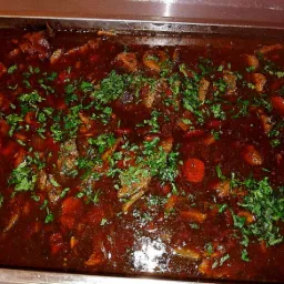 Gulasz wołowy w ciemnym sosie, udekorowany świeżą natką pietruszki, podany w metalowej brytfannie.