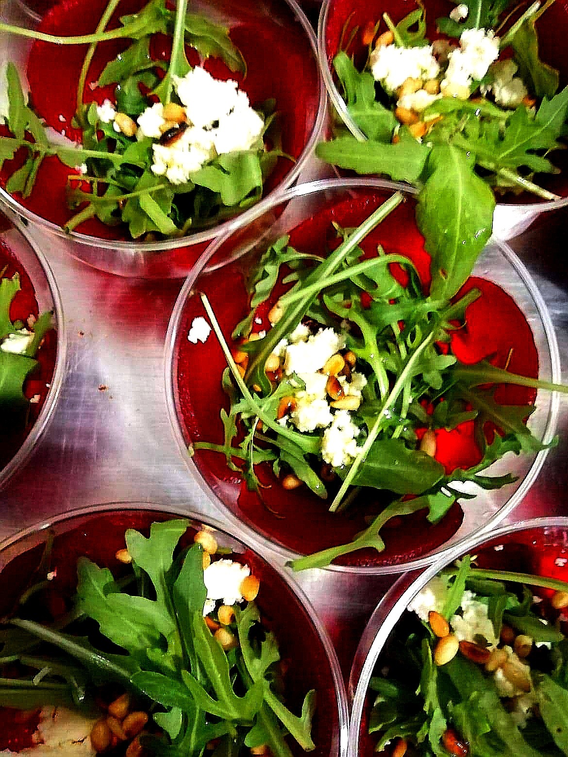 Porcje sałatki z buraka, rukoli, sera feta i orzeszków piniowych w przezroczystych plastikowych miseczkach, ustawione na srebrnej tacy.