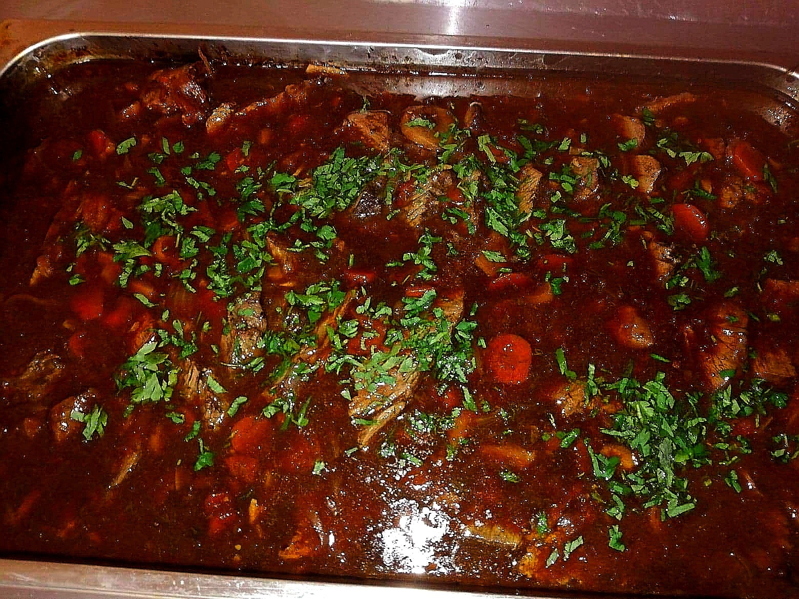 Gulasz wołowy w ciemnym sosie, udekorowany świeżą natką pietruszki, podany w metalowej brytfannie.