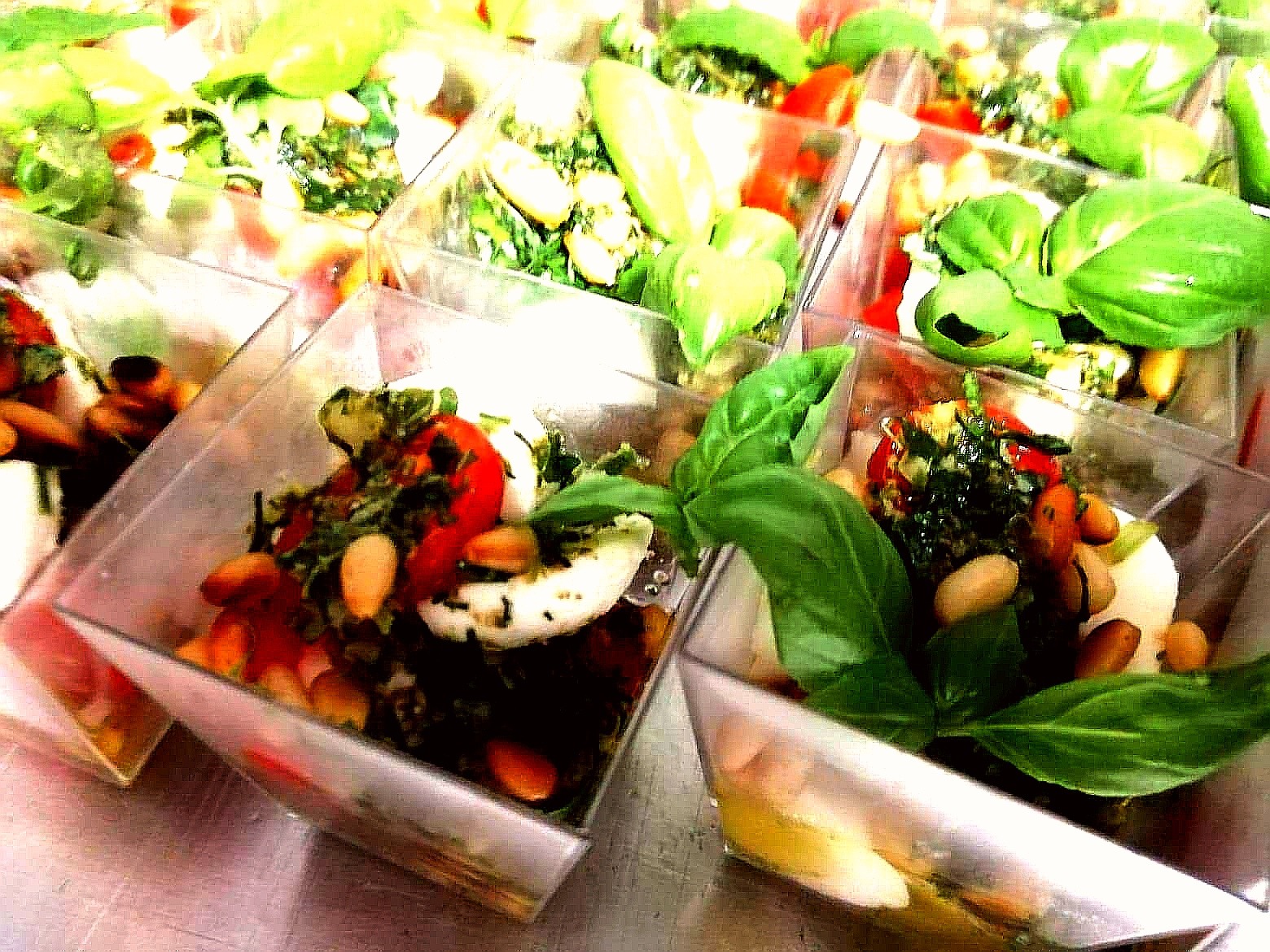 Przystawki w przezroczystych pojemnikach: mozzarella, pomidory, pesto, bazylia i migdały na metalowej powierzchni.