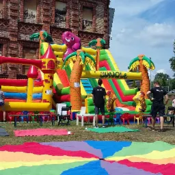 Duży, kolorowy dmuchany zamek z motywem dżungli i słoniem, ustawiony na zewnątrz przed budynkiem z cegły. Wokół rozłożona kolorowa chusta animacyjna, a obok stoją osoby w czarnych koszulkach...