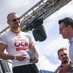 Mężczyzna z bliznami na twarzy w koszulce Dziady OCR trzyma puchar, rozmawiając z dwoma innymi mężczyznami na tle konstrukcji scenicznej.