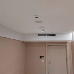 Pomieszczenie z zamontowanym sufitem podwieszanym z wbudowanymi głośnikami, czujnikiem dymu i kratką wentylacyjną, ściany pomalowane na jasny brzoskwiniowy kolor, widoczny fragment drzwi.