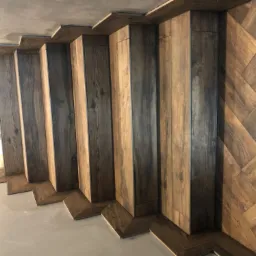 Układanie parkietu w jodełkę na ścianie, widoczne pionowe pasy ciemnego drewna oraz ukośny wzór w jasnych odcieniach, surowy betonowy sufit.