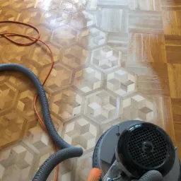 Parkiet o geometrycznym wzorze w trakcie renowacji z użyciem szlifierki; widoczna różnica w kolorze i strukturze między obszarem po obróbce a fragmentem przed cyklinowaniem.