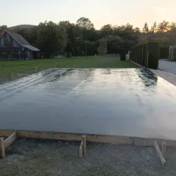 Świeżo wylana, mokra płyta betonowa z drewnianym szalunkiem, odbijająca niebo o zachodzie słońca, z widocznym starym budynkiem gospodarczym w tle.