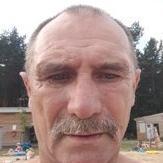 Portret mężczyzny z wąsami na tle plaży, z widocznymi drzewami i budynkiem w oddali. Ujęcie z bliska, skupione na twarzy.