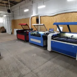 Warsztat z rzędem laserowych ploterów tnących, z otwartymi pokrywami i podłączonymi laptopami, gotowych do pracy.