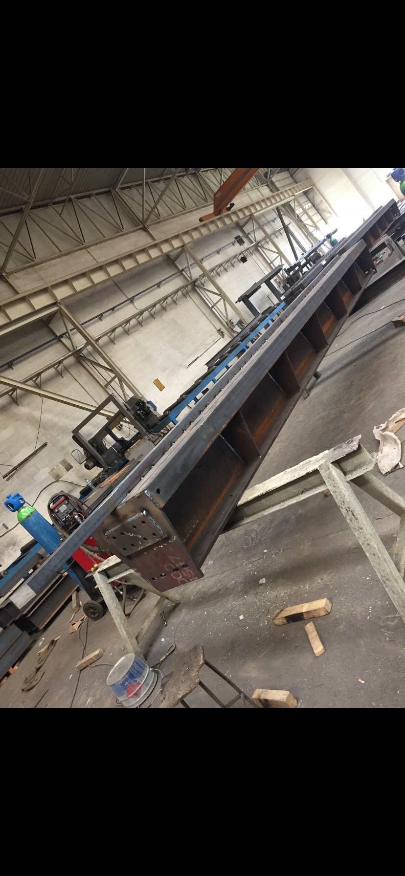 Długa, stalowa belka konstrukcyjna z przyspawanymi wzmocnieniami, leżąca na kozłach w hali produkcyjnej, widoczny sprzęt spawalniczy i suwnica pod stropem.