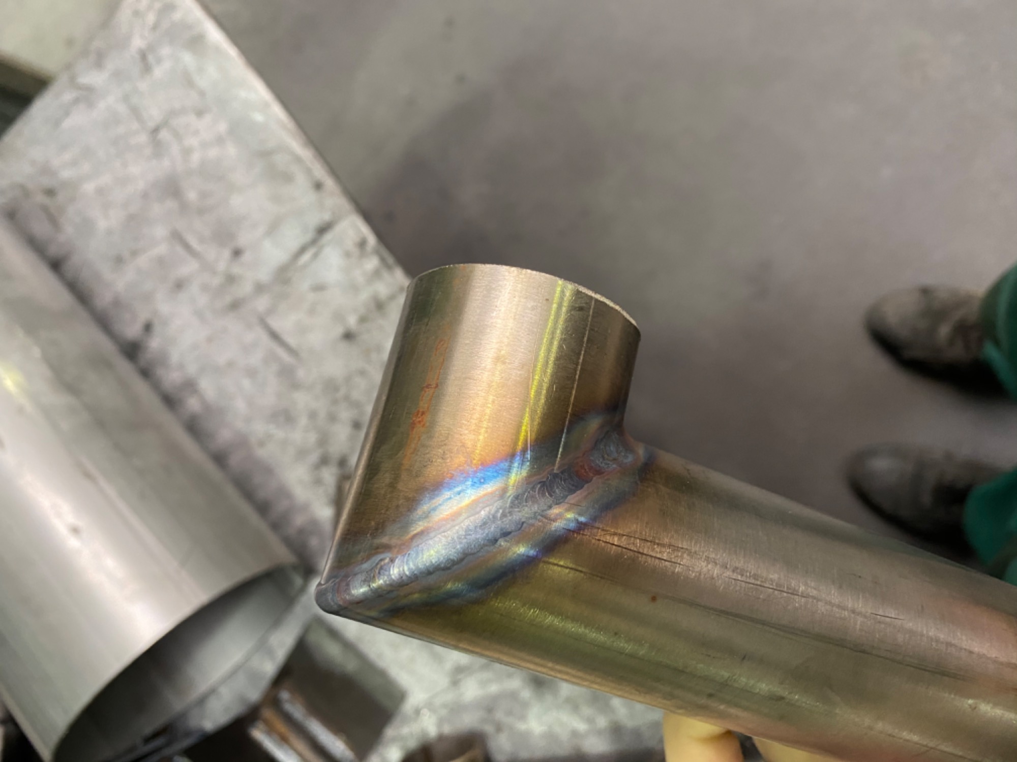 Metalowa rura spawana pod kątem prostym, widoczne ślady spawania z tęczowymi przebarwieniami, szlifowana powierzchnia rury.