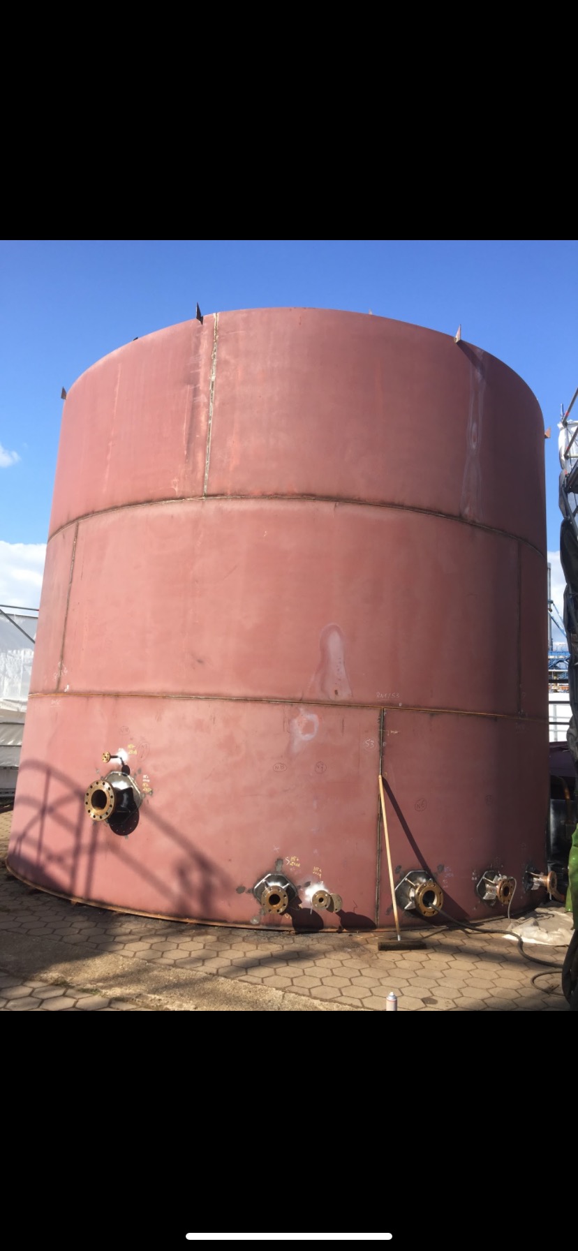 Cylindryczny zbiornik metalowy w trakcie budowy, z widocznymi spawami, przyłączami i oznaczeniami na powierzchni, stojący na kostce brukowej w słoneczny dzień.