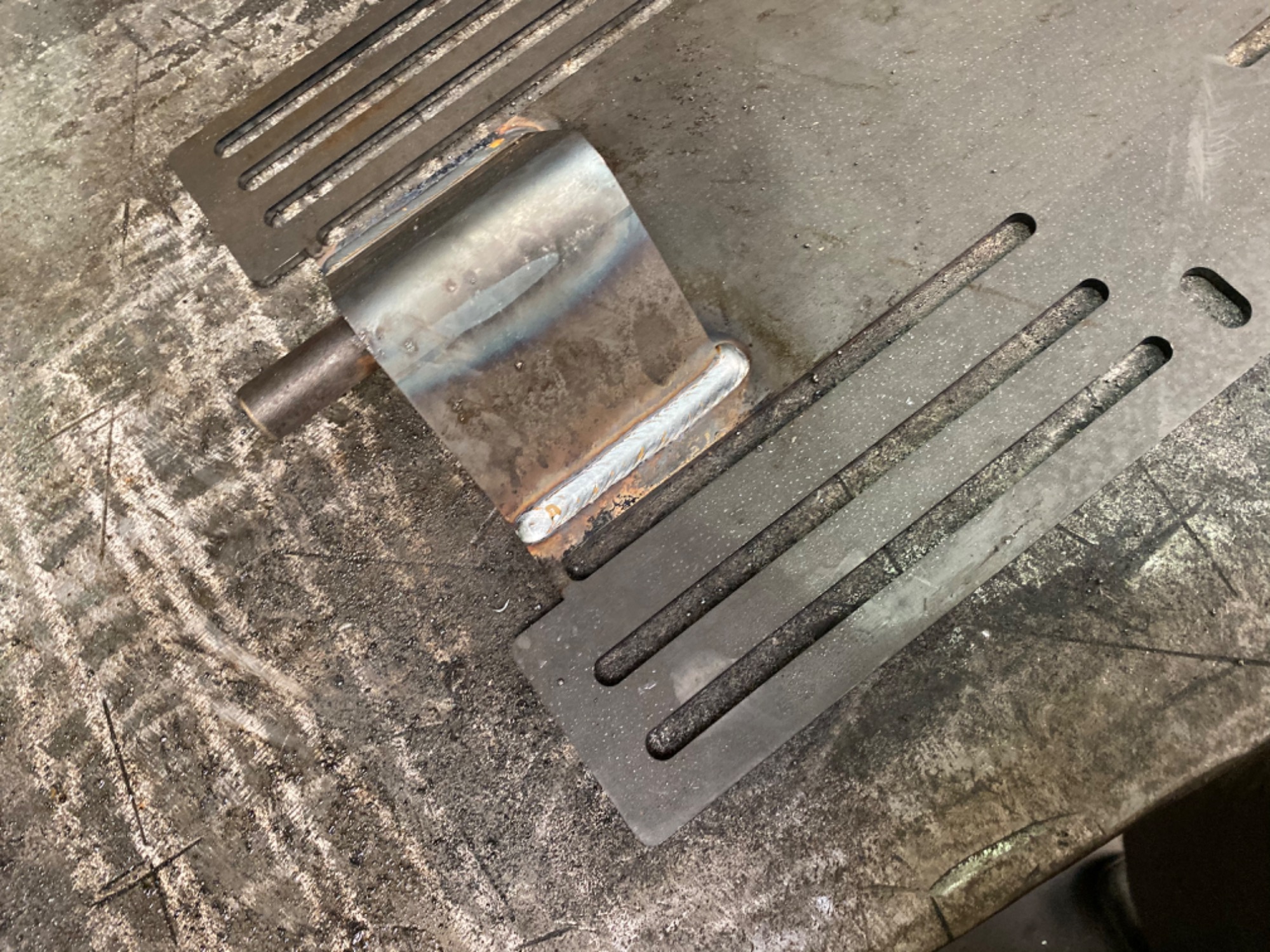 Metalowy element spawany do perforowanej blachy na brudnym stole warsztatowym, widoczne ślady spawania i obróbki.