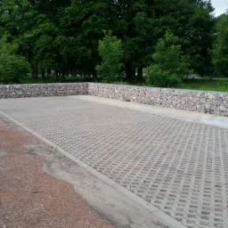 Ukończony parking z ażurowych płyt betonowych, otoczony murem z gabionów wypełnionych kamieniami, na tle zieleni drzew.